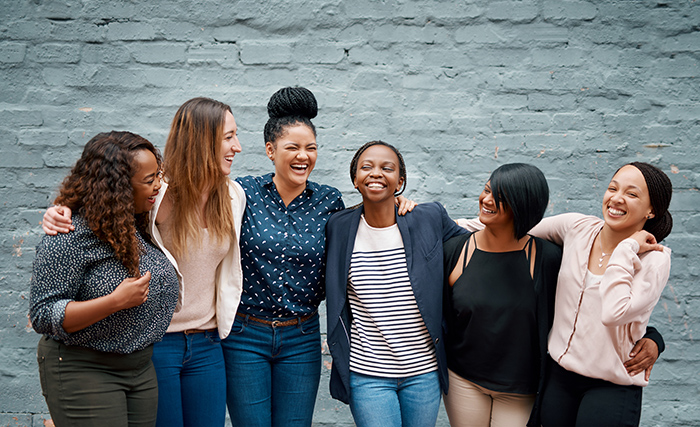 Diverse group of woman with arms around each other smiling in front of gray brick wall