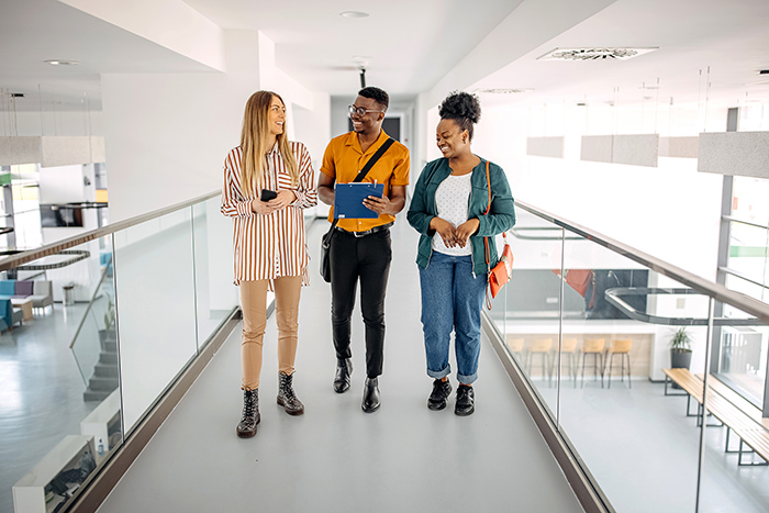 Two women and a man walk along indoor bridge inside office talking