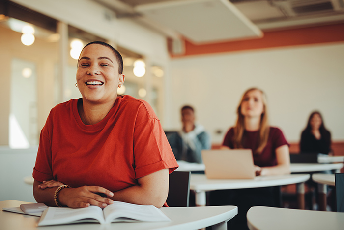 Female college student sitting at desk smiling in classroom with other students in background