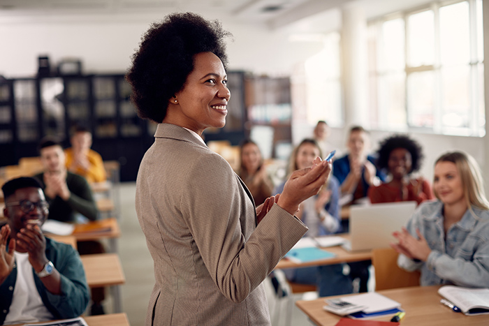 Smiling female professor using pointer in front of class full of students sitting at desks