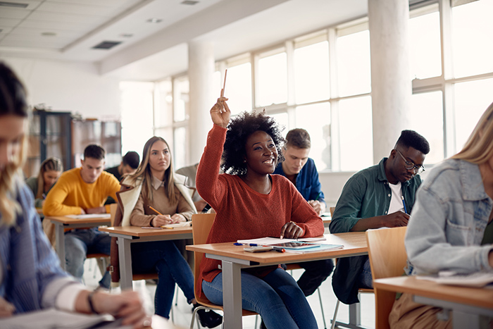 Smiling female college student raising hand in classroom full of peers