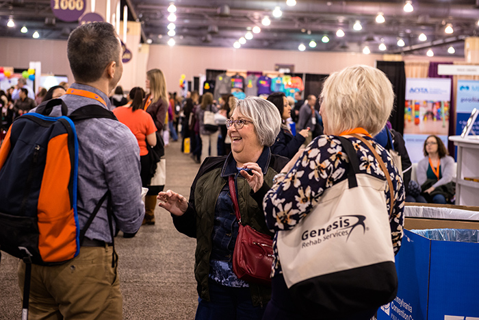 Two female AOTA conference attendees talk to man inside crowded expo hall