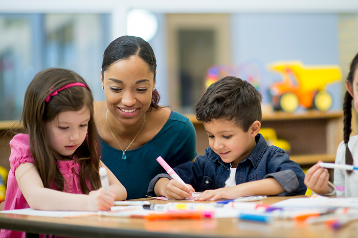 Two small kids holding markers at table with smiling female behind them helping