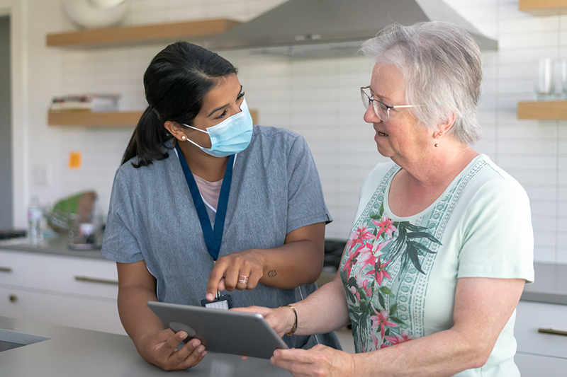 Female occupational therapist wearing a face mask helping senior client in kitchen reading materials on a tablet