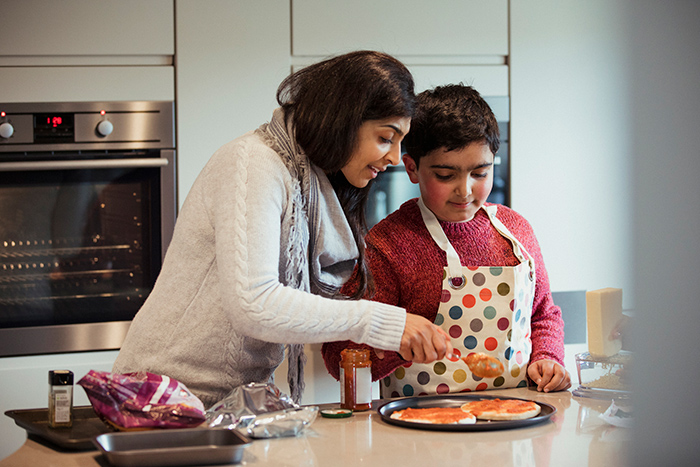 Woman in kitchen helping young boy wearing apron cook pizza