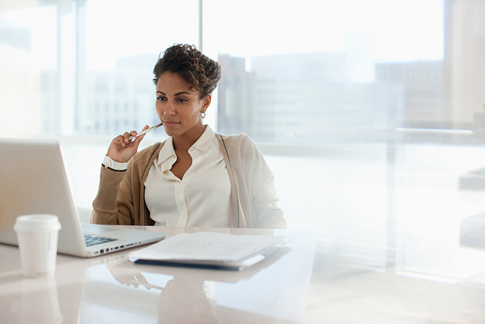 Professional woman sitting at desk reading laptop screen with paperwork beside her
