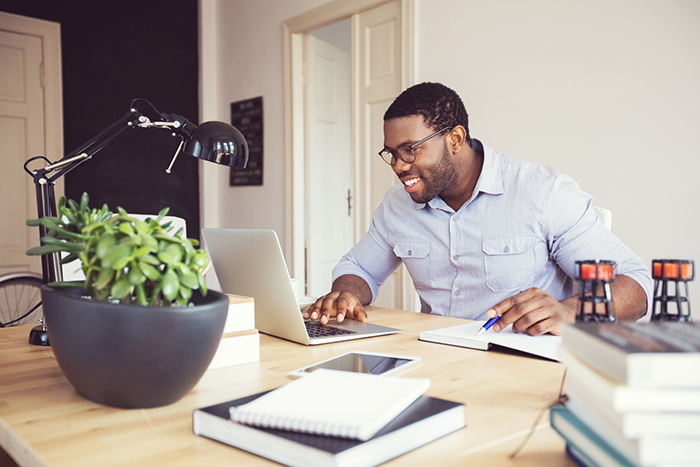 Man smiling at desk looking at laptop and writing in notebook