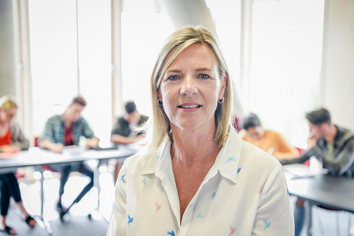 Professional woman looking at camera in front of blurred college students in the background working at tables